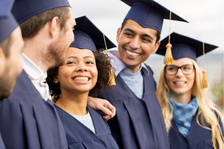 Cinco jovens adultos participando da formatura, todos vestindo becas tradicionais de cor escura e chapéus de formatura com borlas douradas penduradas na lateral.