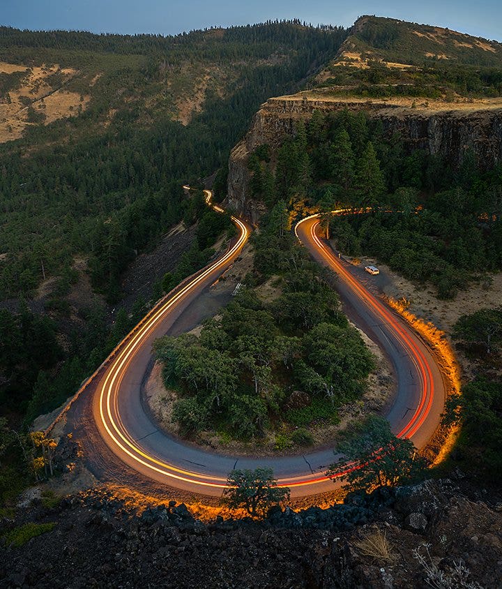 An aerial motion blur photo of cars traveling on a road that runs through a forest hillside