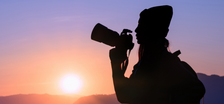 A silhouette of a photographer holding a camera with the sun setting behind them which demonstrates lens photography