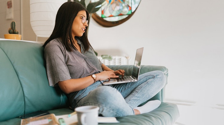 A woman with black hair and glasses on a couch reviewing a purchase agreement on her laptop.