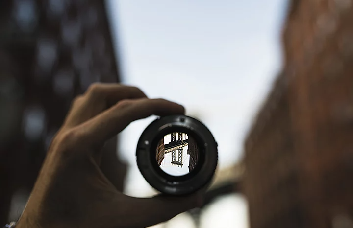 A person holding up a camera lens through which a part of a bridge is showing