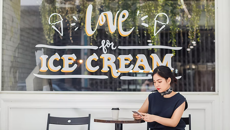 Woman sitting outside in front of a small business that sells ice cream.