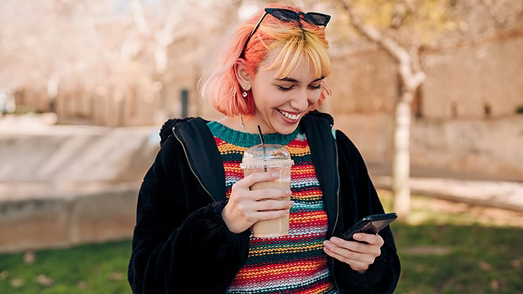A person standing and smiling while looking down at their phone in their hand and holding a coffee in their other hand