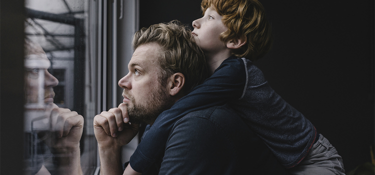 Candid photo of a father with their child piggybacked on them looking out a window pensively