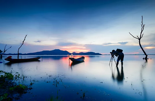 A photographer standing in shallow water capturing the sun setting behind a mountain range