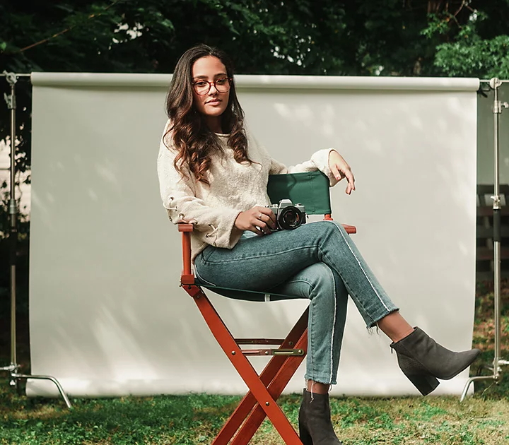 A professional headshot photo of a person sitting in a chair outside with a white backdrop behind them