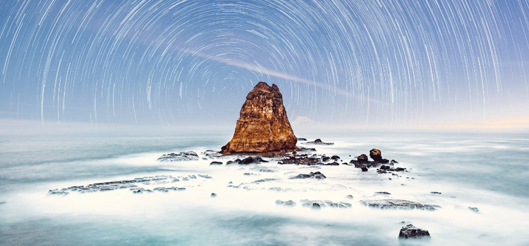 View of ocean with star trails behind it in an example of how long-exposure photography can look