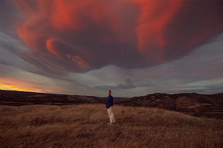 A person standing on grassy terrain with a cloud-covered sky in the background during a sunset taken with a telephoto lens