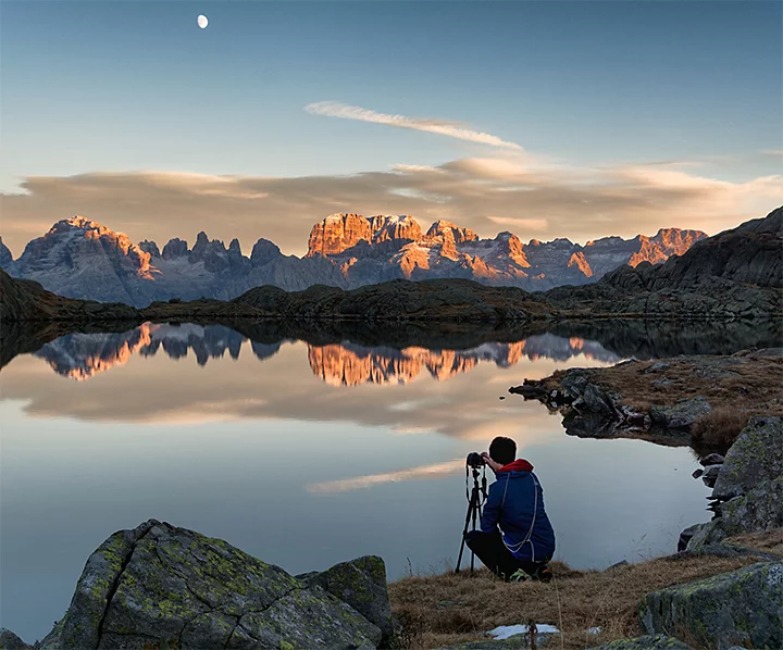 A photographer taking a photo next to a body of water with a rocky mountain ridge in the background