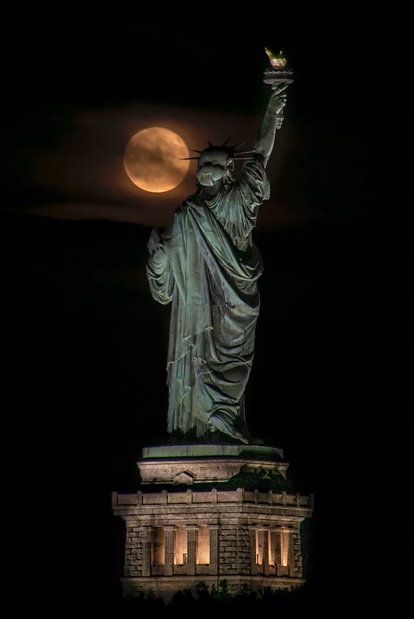 A full moon appearing partially blocked by clouds over the left shoulder of the Statue of Liberty