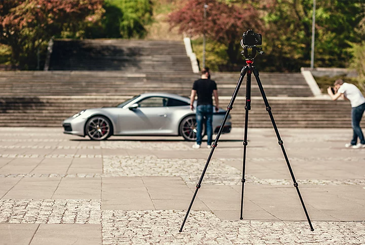 A camera on a tripod with two people and a sports car in front of it