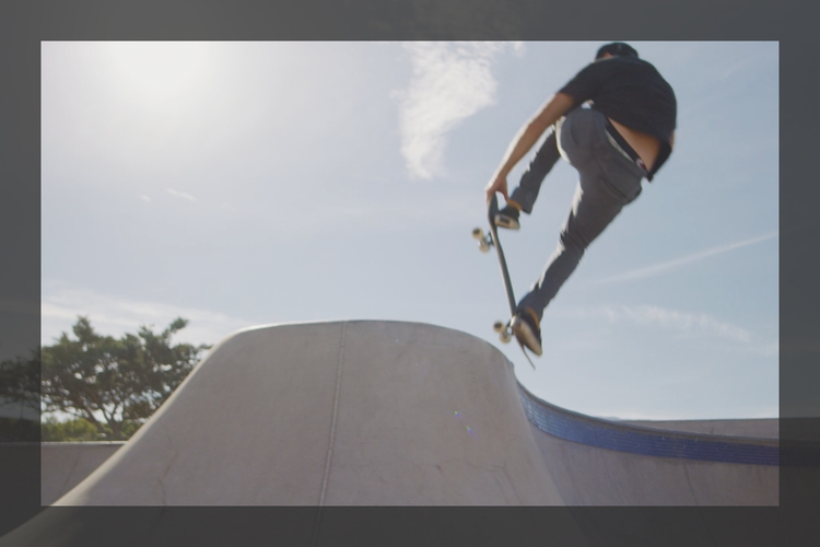 A skateboarder riding into frame and performing a nose grab over the concrete transition of a skate park