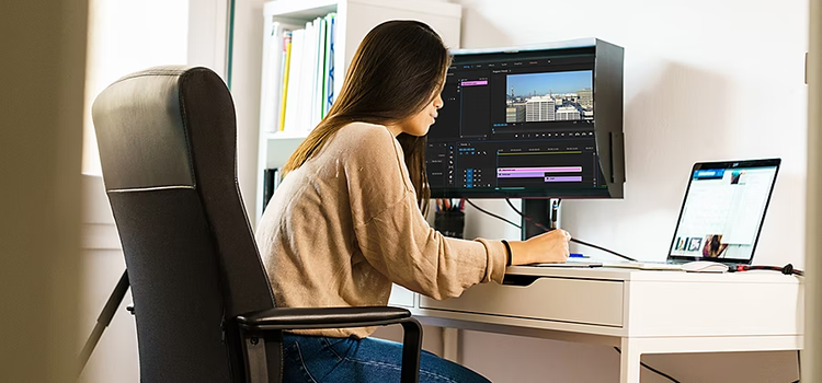 A person sitting at a desk editing a video on their laptop, which is connected to a monitor