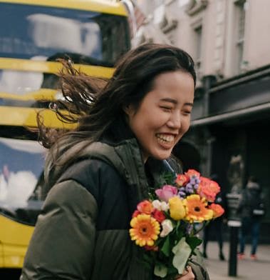 Person carrying flowers being captured in a medium close-up shot in front of a yellow double-decker bus