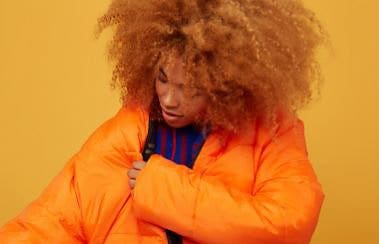 A model pulling on a large orange parka in front of a bold, orange background