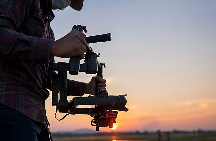 A camera operator uses a handheld DSLR gimbal to film while the fading sun sets in the background.