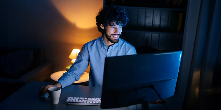 A man in a dark room with a small glowing lamp behind him stands at a desk and smiles at a large external monitor while he compresses a video on his computer.