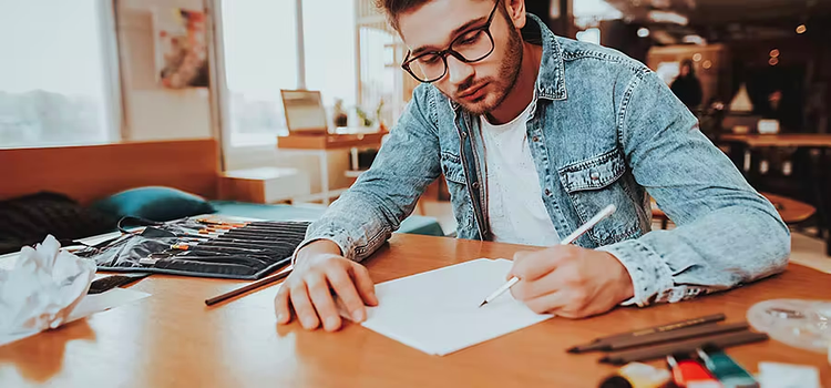 Man sitting at a table with art supplies sketching on a piece of paper.