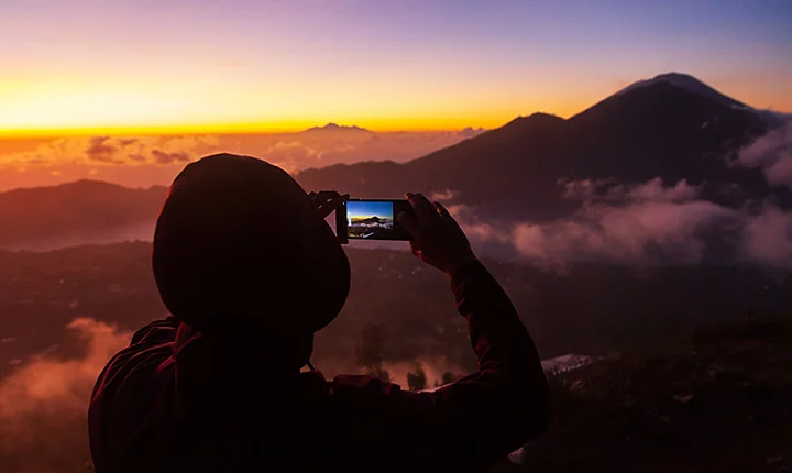 Eine Person, die auf einem Berggipfel steht und ein Foto von einem anderen Berg macht, der sich während eines Sonnenuntergangs durch die Wolken zeigt