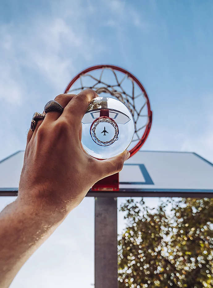 A person holding a lensball up to a basketball hoop while a plane flies high above it