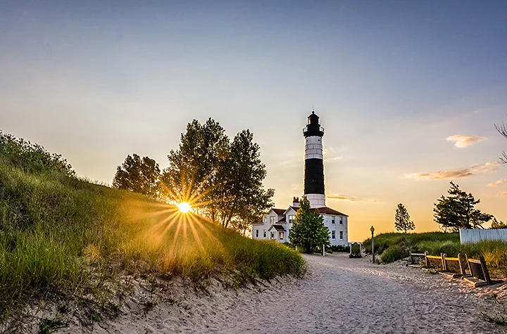 A lighthouse next to a grassy hillside with the sun shining through a tree, creating a lens flare