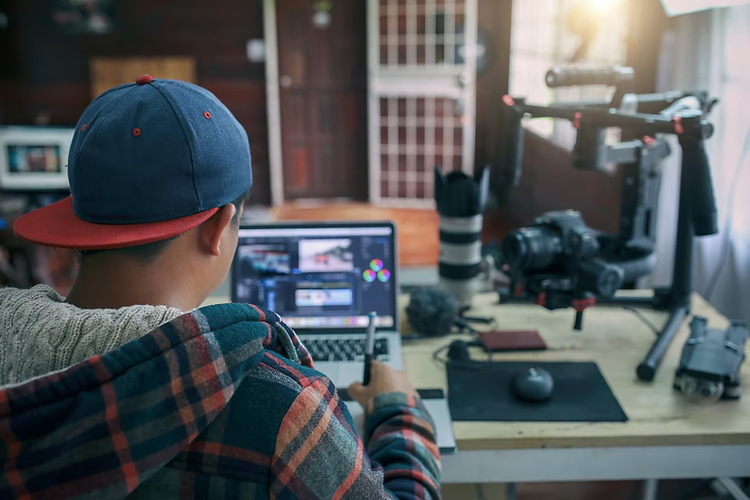 Ein Mann sitzt vor einem Laptop. Er bearbeitet mittels eines Grafiktabletts ein TikTok-Video. Im Hintergrund sind eine Digitalkamera und ein professionelles Zoom-Objektiv sichtbar.