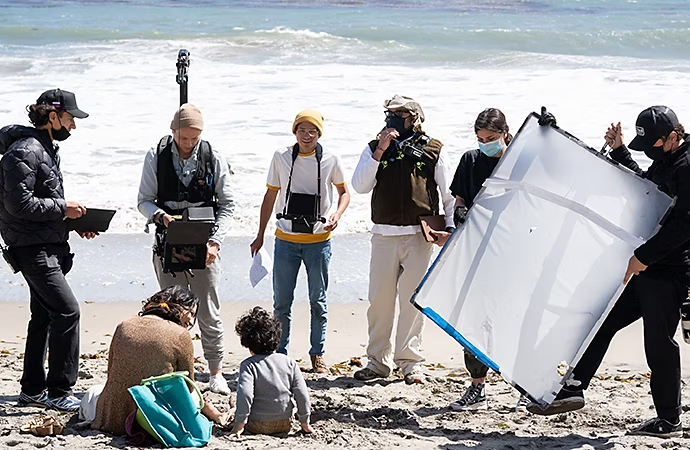 Eine Film-Crew dreht eine Szene, in der eine Person und ihr Kind an einem Strand sitzen.