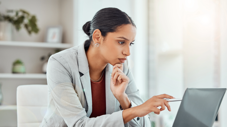 A woman sitting at a desk points looks at a laptop screen and uses a pen to point at the screen.