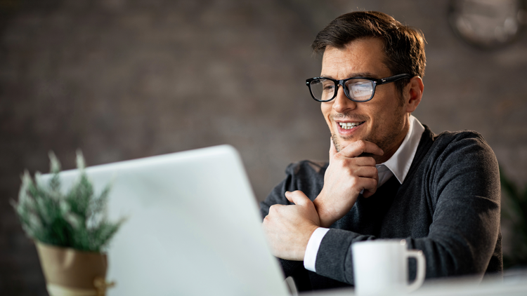 A man looks at a laptop screen and smiles. He is wearing a shirt, jumper, and glasses.