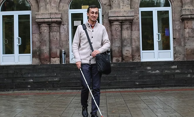 Sipan Asatryan walking in front of building with a shoulder bag and white cane.