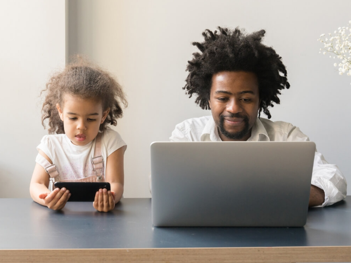 A parent using their laptop and their child looking at their own phone while sitting at a table together