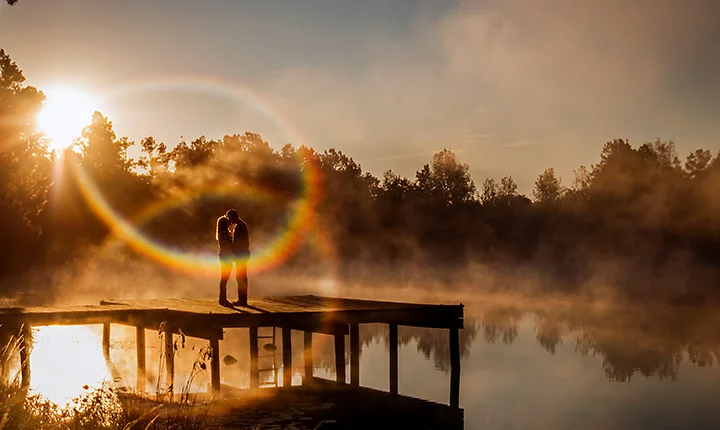 A couple embracing each other while standing on a lakeside dock with a lens flare above them