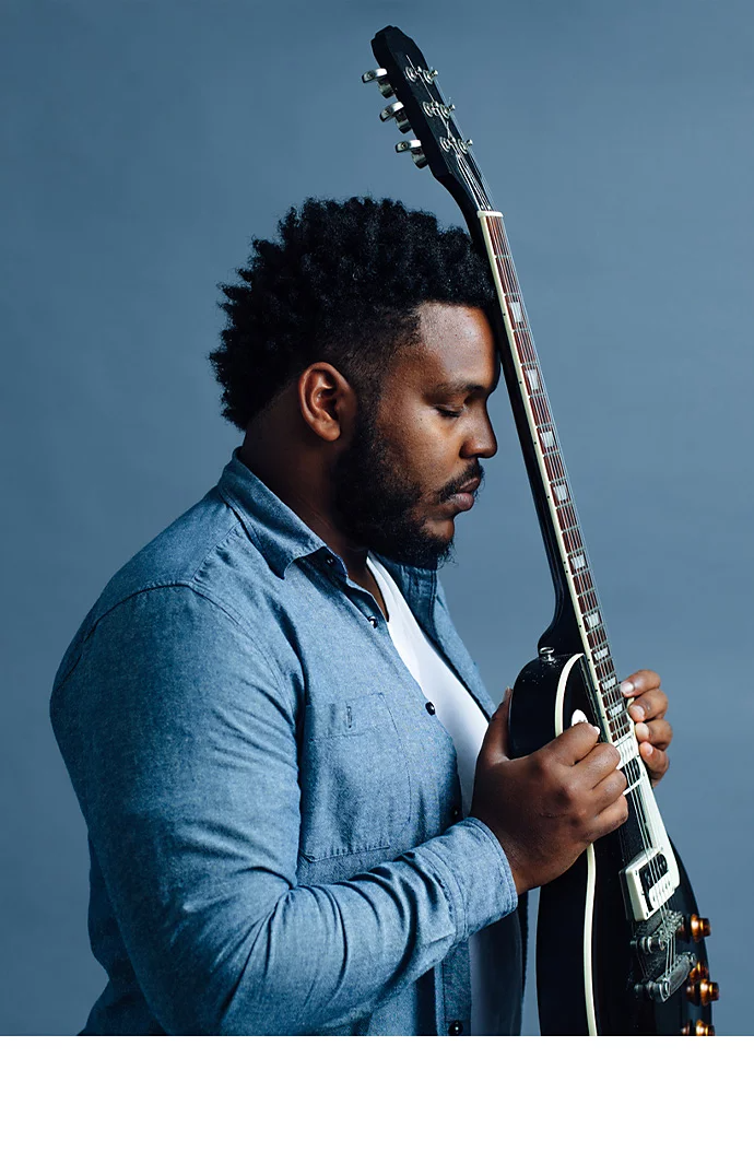 A professional headshot photo of a person holding a guitar in front of their face