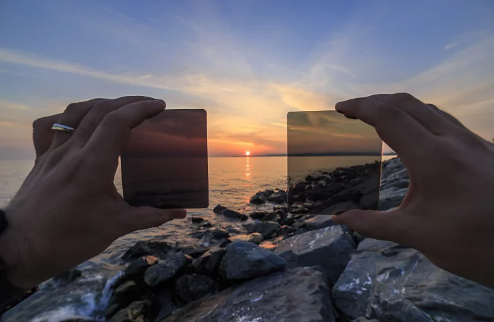 A person holding up two different ND filters to a sunrise