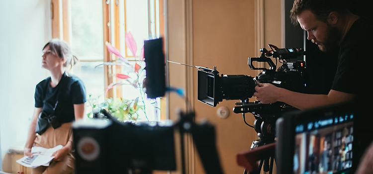 Un plató de producción con un videógrafo mirando por el visor de su cámara y una directora de pie al fondo sujetando un guion