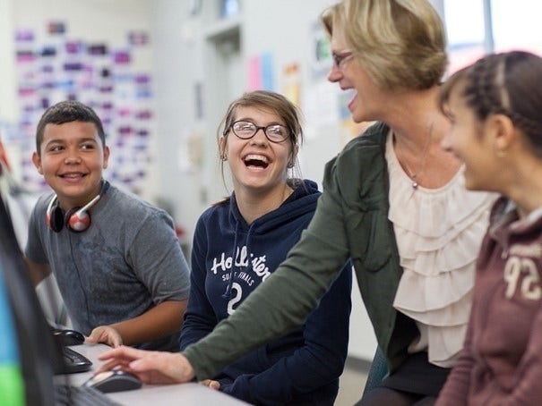 Students sitting at a table in a classroom and a teacher assisting them and using a computer.