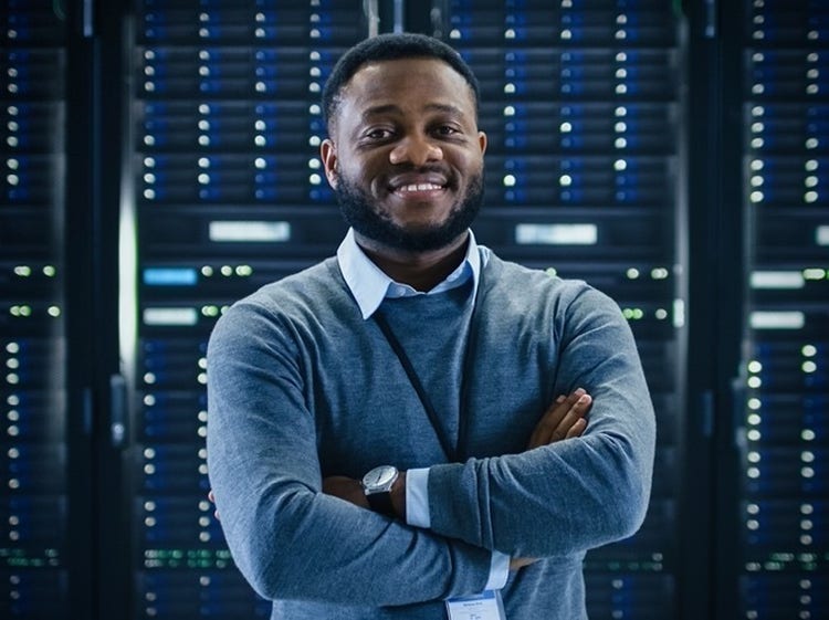 A person smiling with crossed arms standing in front of a server room.