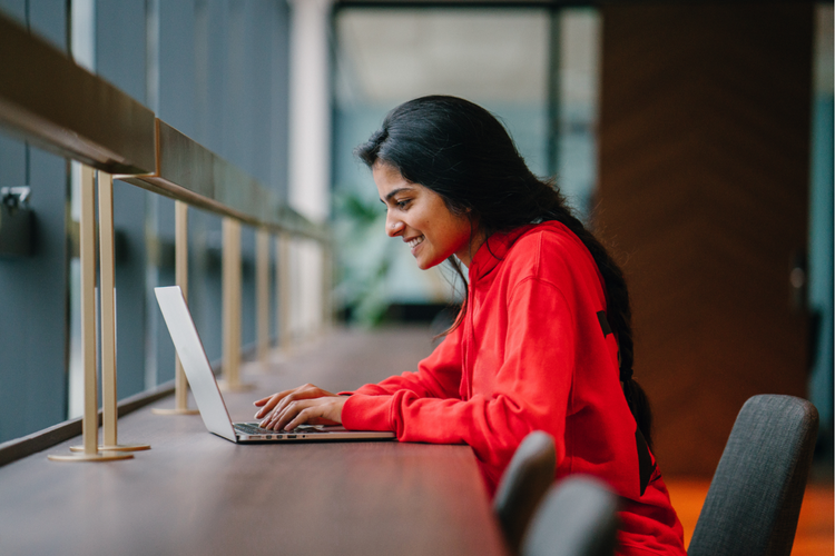 A woman with long dark hair wearing a red blouse sits at a long dark hardwood table working on a silver laptop.