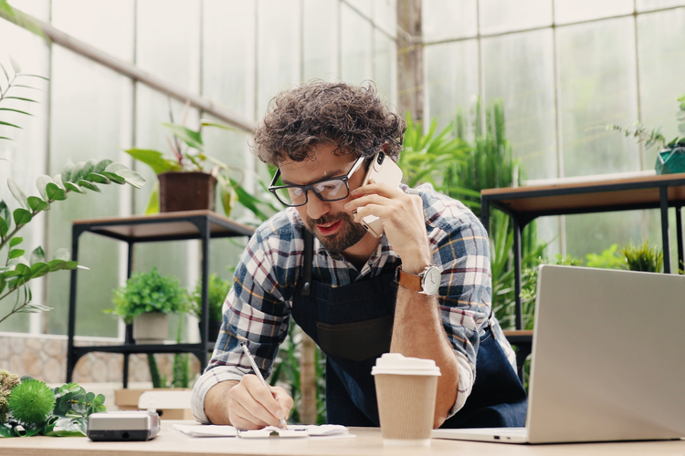 A small business owner on their phone organizing social media marketing while bending over a desk with a notepad, pen, laptop, and a cup of coffee