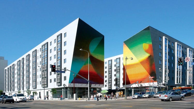 A building at a city intersection that features a colorful digital image split across two surfaces of the building. The image, whic is split into different sections that are colored differently primarily includes a bowl and a finger resting on it, suggesting a hand outside the image.