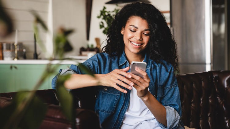 A woman look at her phone and smiles. She is wearing a denim shirt and has dark hair.