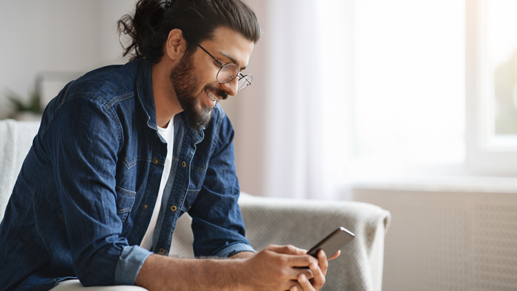 A man sits on a sofa and looks at his phone. He is wearing a denim shirt and glasses.