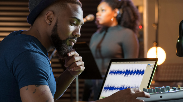 Man in recording studio A man in a recording studio sits in front of a laptop with sound waves on the screen. A woman with a microphone is in the background.