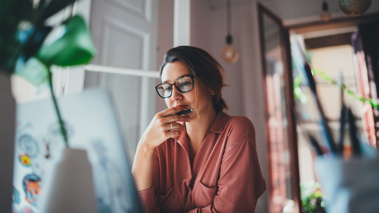 A woman looks thoughtfully at her laptop. She is holding a pen.