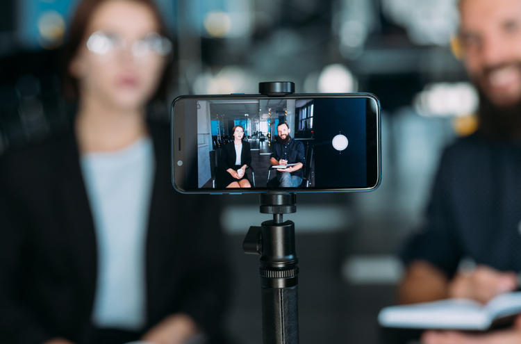 A phone set up sideways on a stand recording a YouTube video of two people sitting in chairs