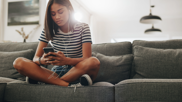 A young woman sits cross-legged on a sofa and looks at her phone. She is wearing wired earphones.