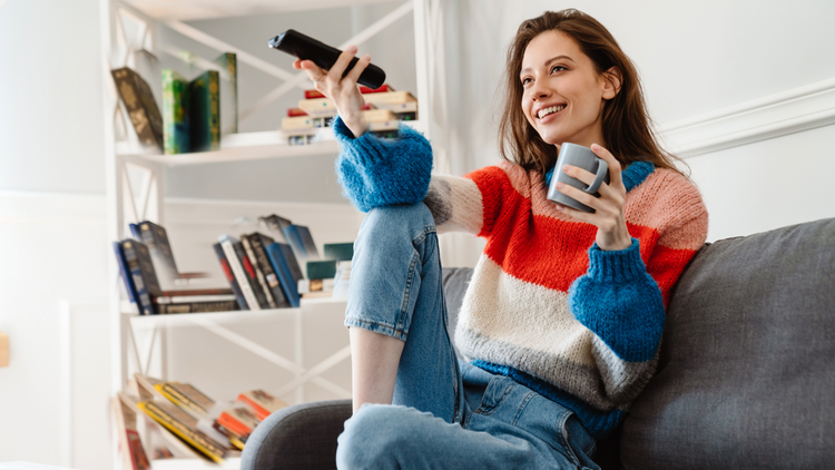 A woman sits on a sofa with a television remove in one hand, and a mug in the other.