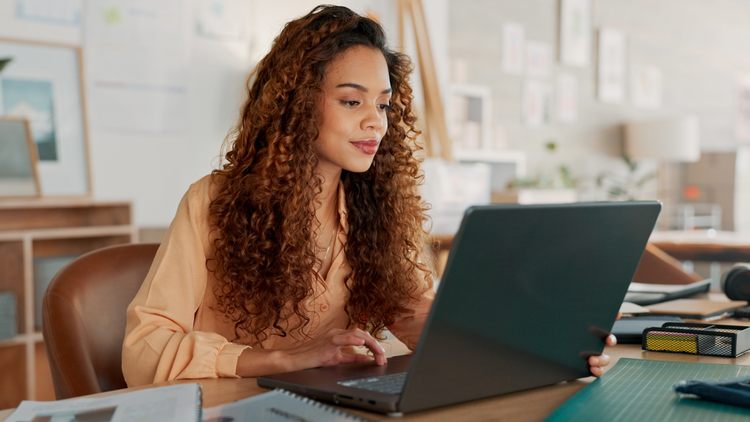 A woman sits at a desk with her laptop. She looks at the screen. She is wearing a peach-colored blouse.