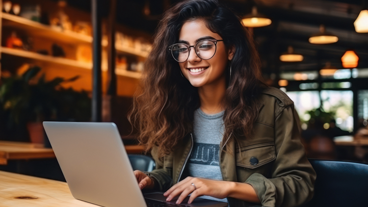 A woman sits at a table with her laptop in front of her. She looks at the screen and smiles. She is wearing glasses.