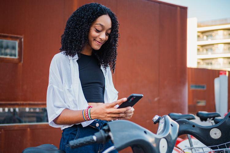 A young woman stands outside looking down at her phone. She is smiling.